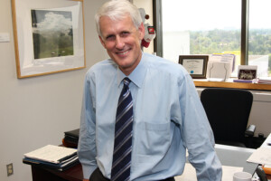 A man wearing a dress shirt and tie leans against a desk while posing for a photo.