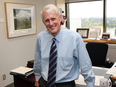 A man wearing a dress shirt and tie leans against a desk while posing for a photo.