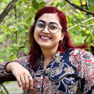 Meral is outside leaning on a tree branch, with red long hair, glasses and a multi-coloured blouse