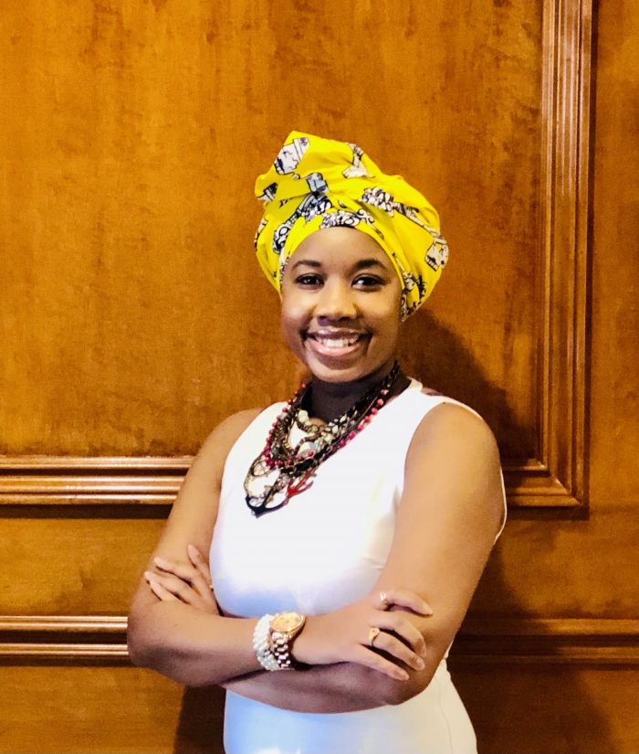 Rumbi is standing by a wooden wall, wearing a yellow & black head scarf, brown & red necklaces & a white sleeveless top