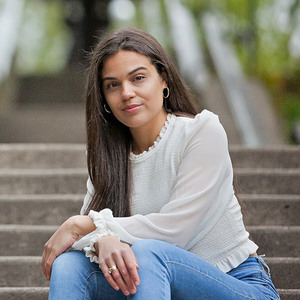 Samira is sitting outside on steps, has long brown hair, white blouse and jeans.