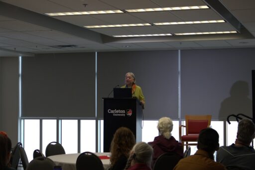 Elder Barbara in a green sweater, standing at a podium speaking to conference delegates.