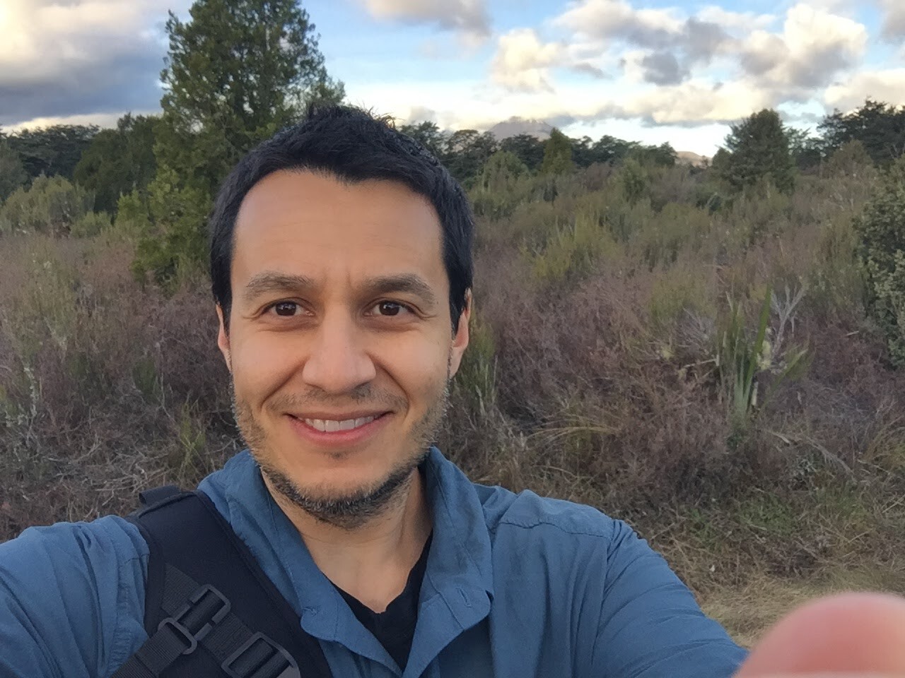 Man with dark short hair standing in a field, wearing a blue shirt, crossover black bag and smiling - taking a selfie. 