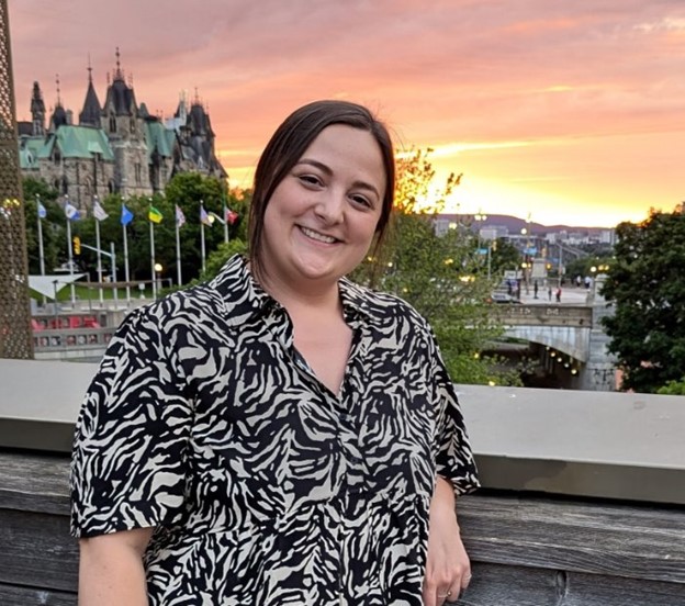 Woman standing outside near Parliament Hill, smiling, with black hair tied back wearing a black and white blouse. 