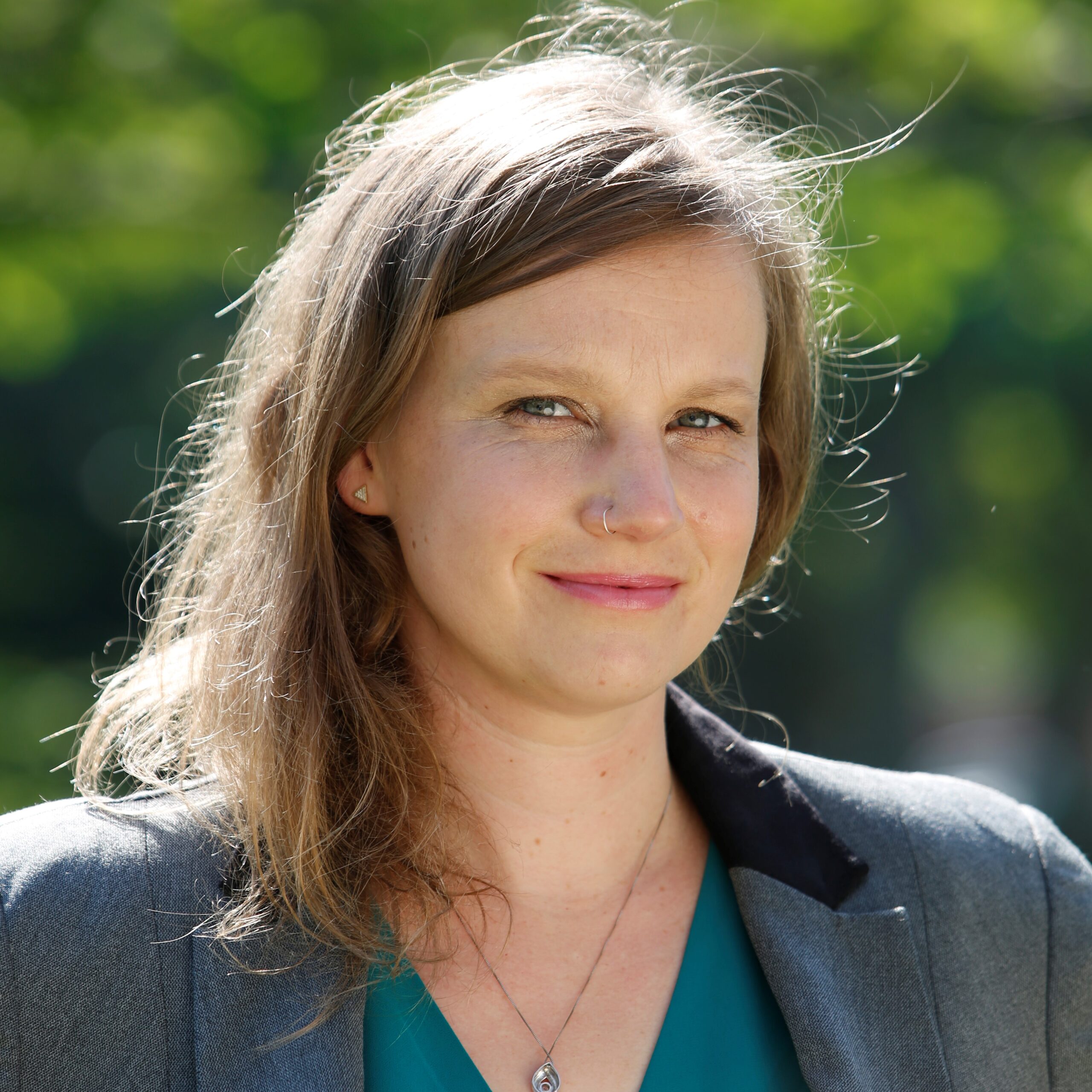 Woman with brown long hair, lipstick, grey blazer and blue blouse standing outside in front of trees. 