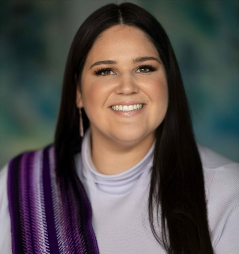 Woman with long black hair and a smile wearing a light purple top with a dark purple and white wool scarf on her shoulder. 