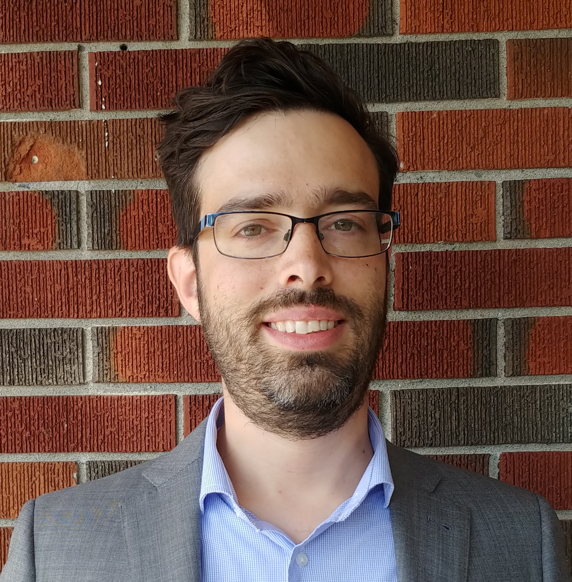 Man with short black hair and beard standing in front of a read brick wall wearing a grey blazer and light blue shirt. 