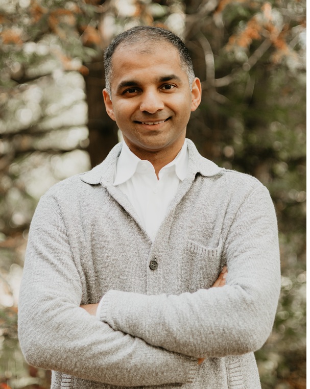 Man with short salt and pepper hair, standing outside wearing a grey cardigan and white shirt, smiling. 