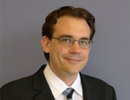 Man with dark hair and glasses in a black suit jacket, white shirt and black tie. 