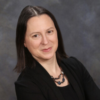 Woman with shoulder length black hair smiling wearing a black blazer, top and beaded necklance. 