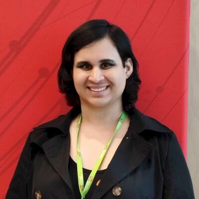 Black shoulder length hair,smiling and standing in front of a red backdrop wearing a black top and a neon green lanyard. 
