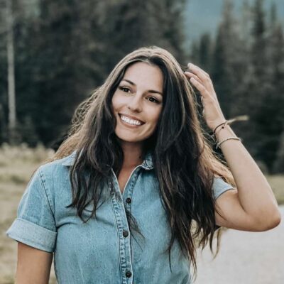 Standing outside with a forest behind her. She has dark brown long hair, is smiling and wearing a short sleeved jean shirt
