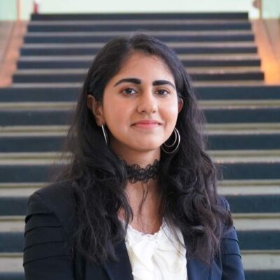 Standing at the bottom of stairs, with black, long hair, hoop earrings, a black blazer and white blouse. 
