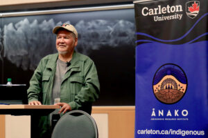Wilfred Buck delivers a talk. He is smiling at the audience and next to him is a banner advertising the Anako Indigenous Research Institute.
