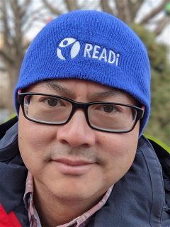 Adrian Chan is wearing his blue beanie outside. He is looking at the camera.