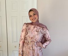 Farah is standing in front of a white door, smiling at the camera. She is wearing a pink dress and a dark pink hijab.