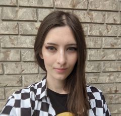 Georgia is standing in front of a brick wall, looking ath the camera nad wearing a checkered shirt.