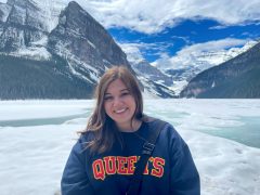 Mackenzie is standing in front of snow capped mountains and smiling at the camera. She is wearing a Queen's sweatshirt.