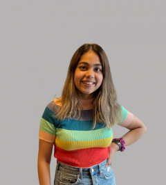 Manya is smiling at the camera in front of a white wall. She is wearing a t-shirt with rainbow stripes.