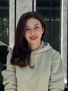 Shaghayegh is looking at the camera in front of a building. She is wearing red lipstick and a sweatshirt.