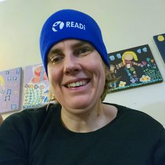 A woman is wearing a READi blue beanie and smiling. Behind her is a wall with colourful pictures.