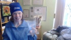 Glenda Watson Hyatt is in her home office, wearing a READi blue beanie. She is smiling and holding a Mitacs award. Next to her is a sleeping grey cat.