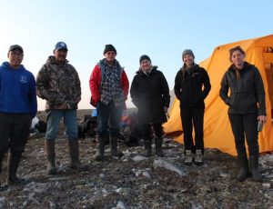 Three men and three women stand in the Arctic with camping gear and a yellow tent in the background.