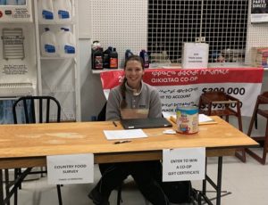 A young woman sits at a wooden table in a grocery store ready to conduct surveys.