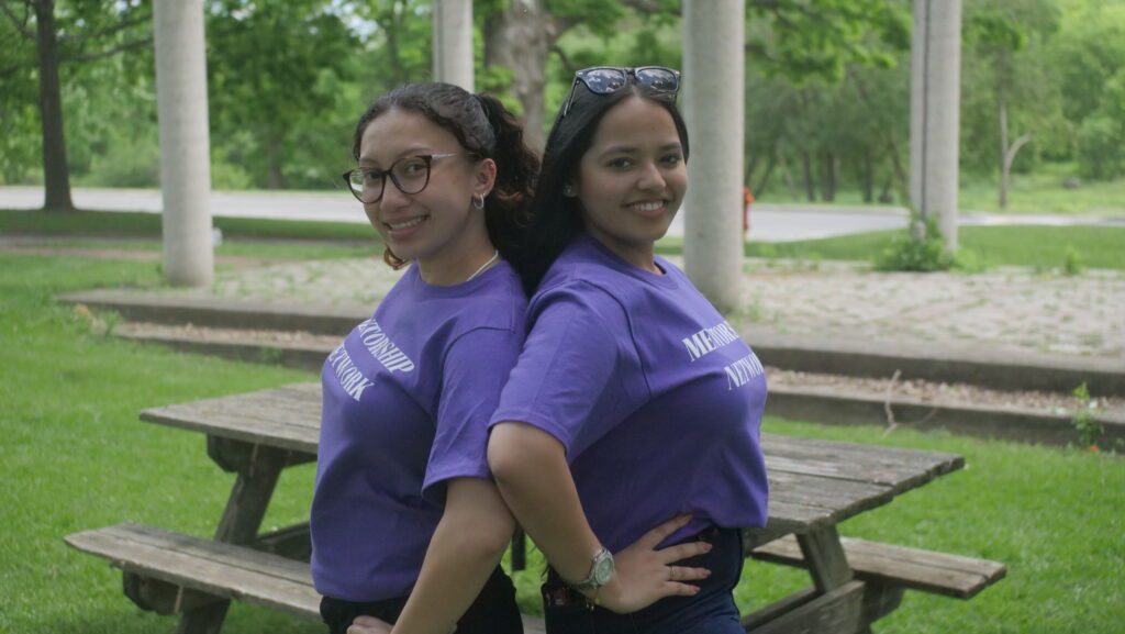 Two smiling First Year Connections Mentors wearing purple shirts stand back to back in front of a picnic table outside.