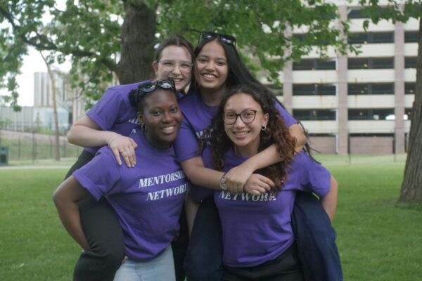 Four smiling First Year Connections Mentors wearing purple shirts outside.