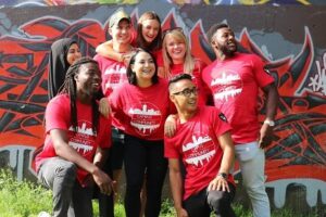 Group of students with their campus to community t-shirts posing for a group photo in front of a wall of graffiti.