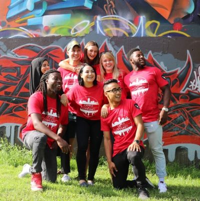Group of students with their campus to community t-shirts posing for a group photo in front of a wall of graffiti.