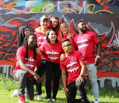 Group of students with their campus to community t-shirts posing for a group photo in front of a wall of graffiti.