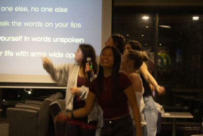 Group of students singing and dancing to karaoke with lyrics on screen behind them.