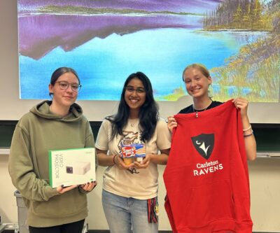 Three students showing off their raffle prizes: Video Projector, a package of pudding snacks, a Ravens sweater.