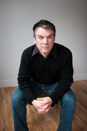 A picture of Michael de Adder sitting in an empty room. de Adder is a white man with short grey hair, and he wears a black dress shirt and blue jeans.