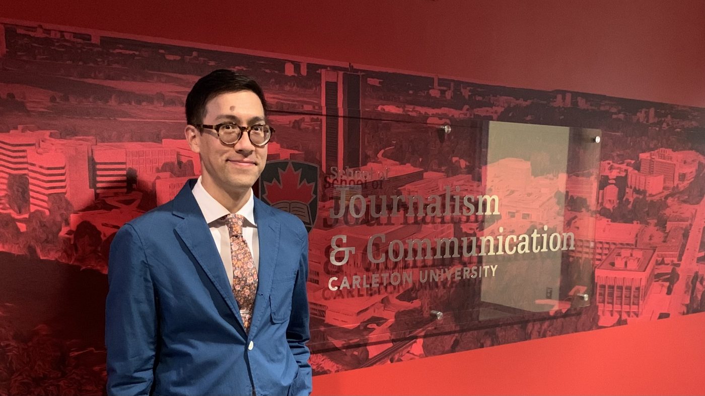 Prof. Benjamin Woo stands in front of a red wall that is printed with an aerial photograph of Carleton campus overlaid with a glass sign reading "School of Journalism & Communication, Carleton University."