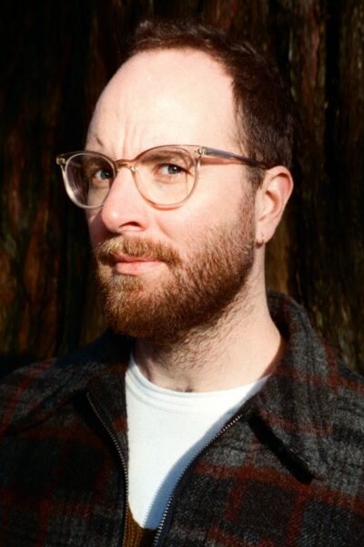 A portrait of Dylan Mulvin, a white man with short brown hair, a short brown beard, and glasses. He wears a dark plaid jacket over a white t-shirt and is looking at the camera with a serious expression..