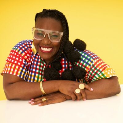 Nana aba Duncan, a Black woman with glasses and braids ending in puffs of hair, smiles in front of a yellow background. She is wearing gold jewelry and a multi-coloured checked shirt.