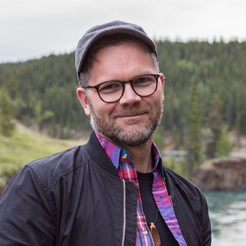 A headshot photo of Matthew Pearson, a white man with a short beard and glasses. He is wearing a plaid shirt, a black bomber jacket, and a black baseball cap, and stands in front of a tree-covered mountain.