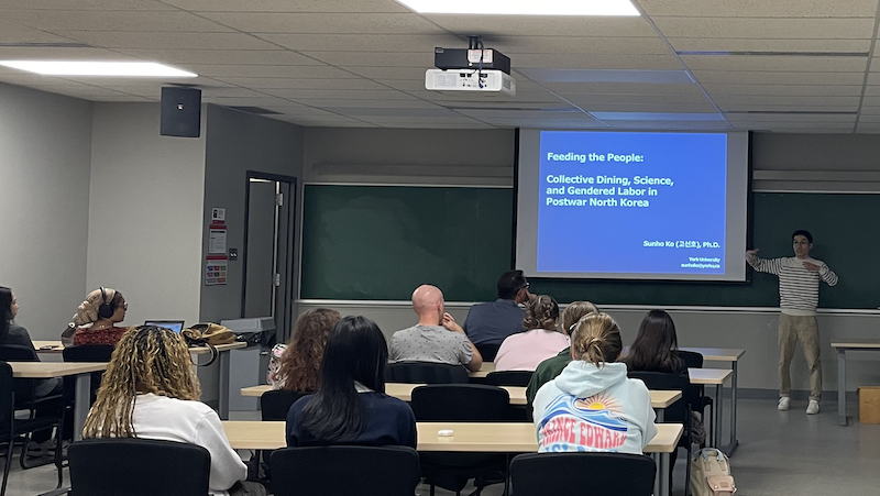 Dr. Ko presents to a room of students. We see the screen from the back of the room. Slide reads: "Feeding the People: Collective dining, science, and gendered labor in Postwar North Korea."