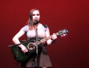 Emily plays her guitar onstage with a red background