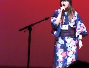 Aki dressed in a flowered kimono stands and sings onstage with a red background