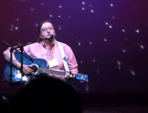 Euan in his pink shirt sits and plays his blue guitar against a backdrop of purple sky with stars