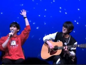 Joe waves to the crowd while Ruoxin plays the guitar with a blue background and stars