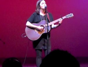 Sarah plays her guitar and sings while standing on the stage with a red background