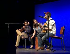 Gythe, Mélissa, and Arno seated on the stage. Mélissa is sitting on and playing a cajon. Gythe and Arno are playing guitars.