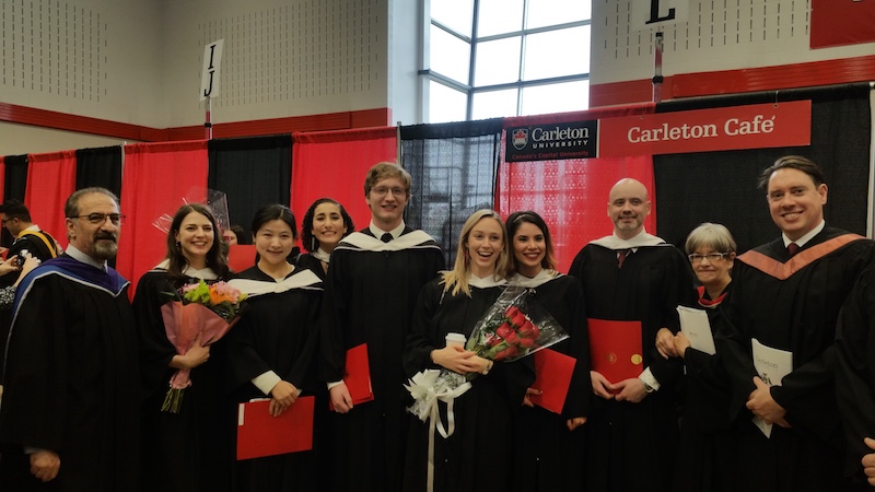 MA grads stand in front of Carleton banners holding diplomas