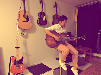 Yang playing his guitar against a wall with guitars hung on it.