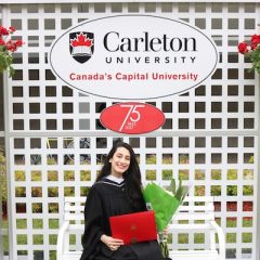 Raghad seated in front of the Carleton University sign holding her degree and a bouquet of flowers.